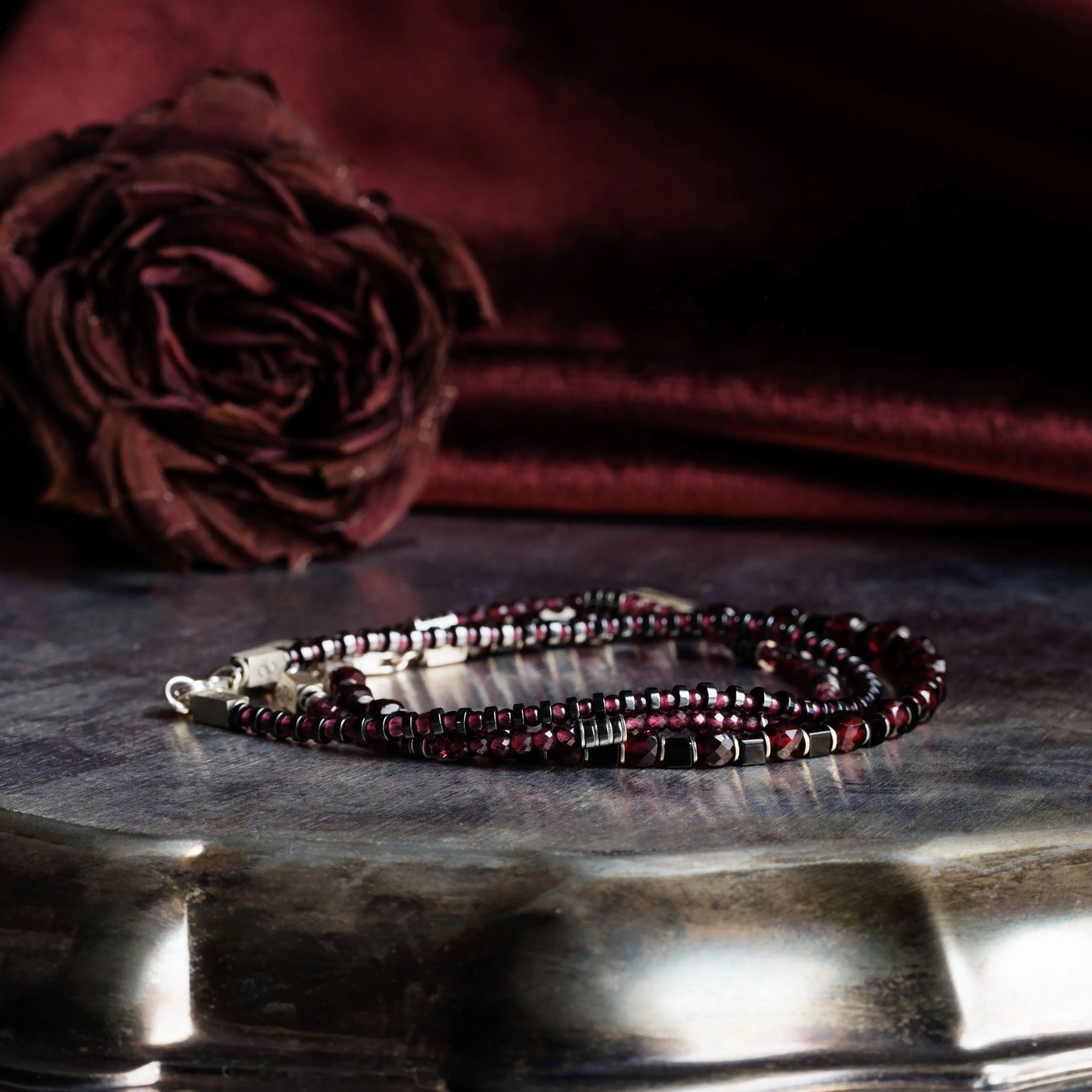 Elegant Red Garnet Bracelet XIII with 2mm stones and silver lock, styled with a dark rose and luxurious fabric backdrop.